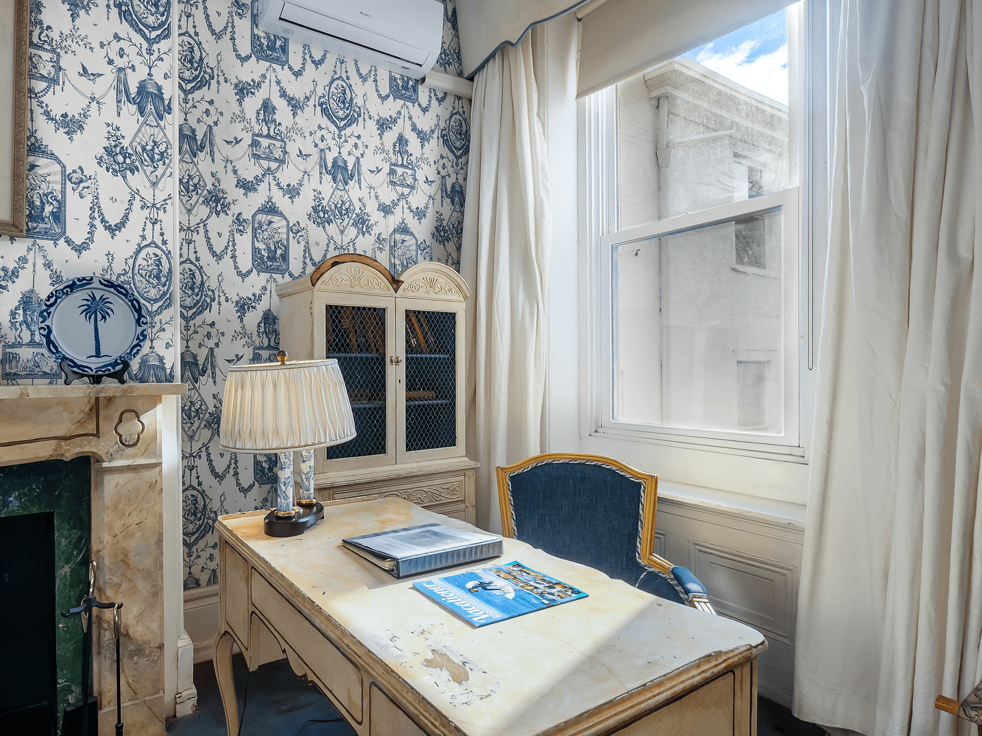 A bright room with blue floral wallpaper, a wooden desk, a lamp, and a window.