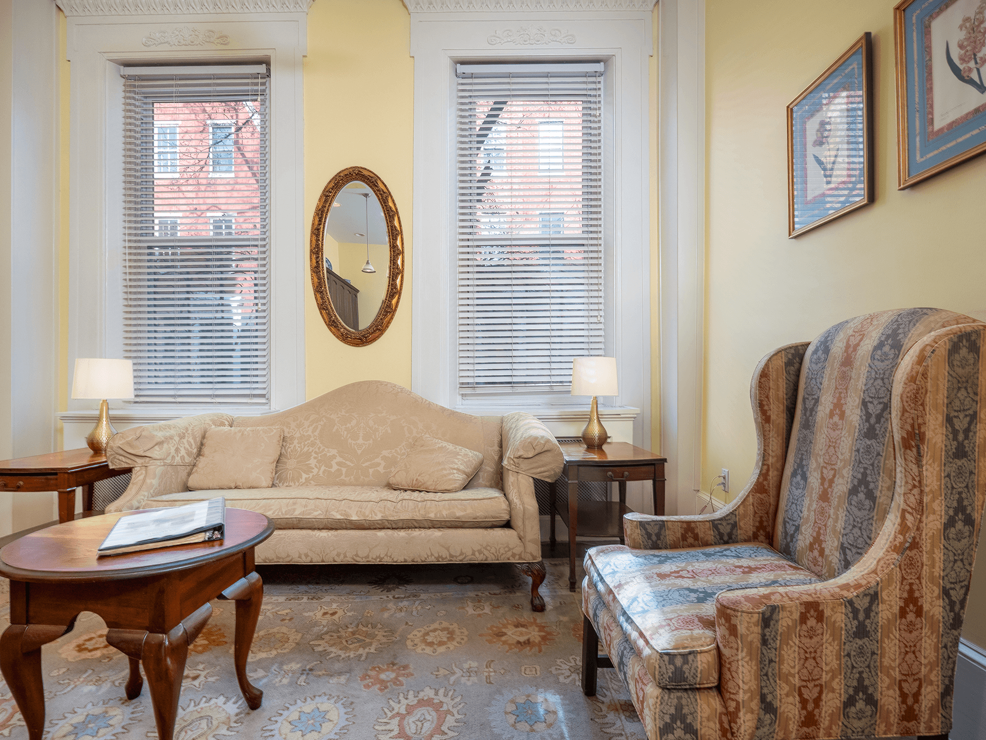 A cozy living room featuring a beige couch, a patterned armchair, a wooden coffee table, and two windows with natural light.