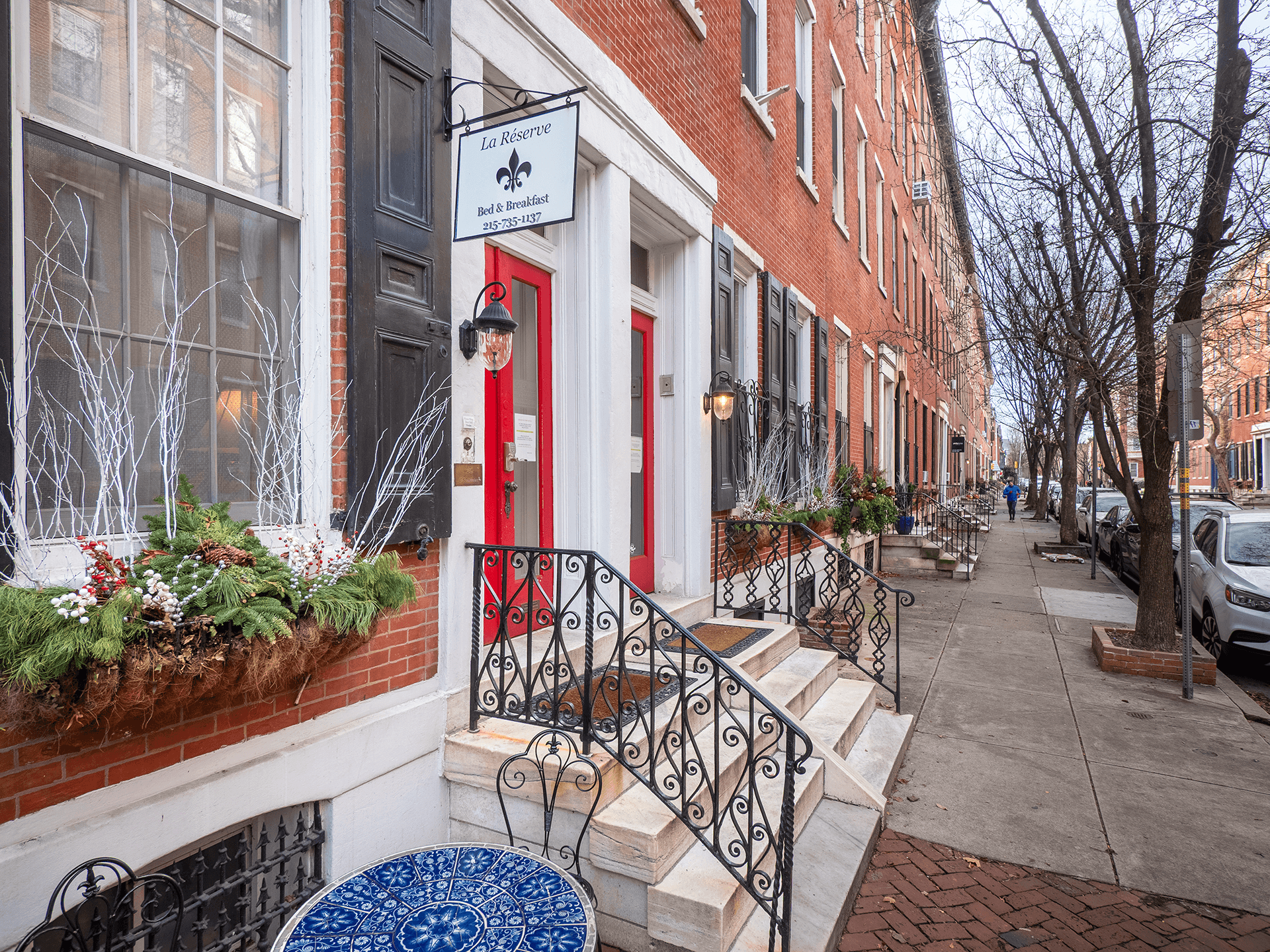Entrance of a red brick bed and breakfast with decorative planters and a blue table on the sidewalk.